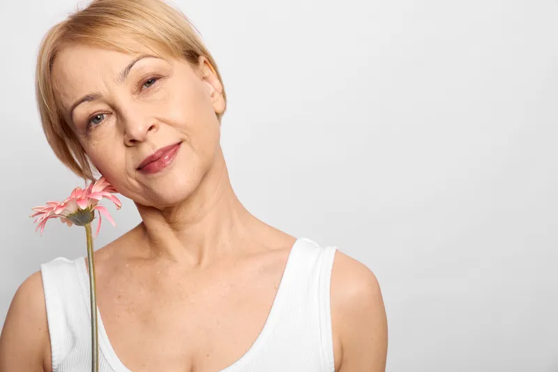 Woman portrait in studio, closeup on face, holding pink flower, gentle smile, bare shoulders, soft light on white background, relaxed calm mood, natural makeup skincare