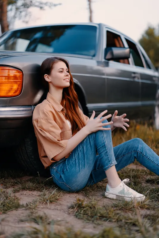 Young woman in casual outfit sitting by a car, expressing relaxation and joy in a natural setting during sunset. Casual, outdoor, happiness, lifestyle.