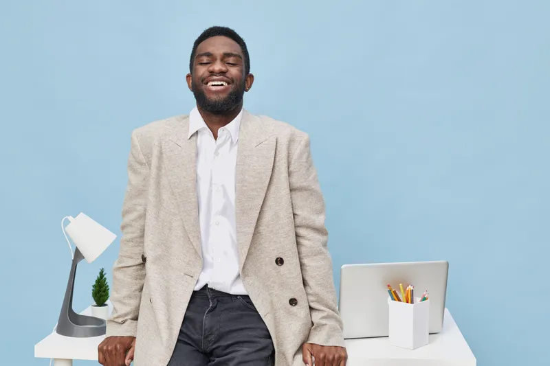 Smiling man in beige blazer and white shirt standing beside modern office desk with laptop, stationery, and plant, cheerful expression, professional appearance, bright background, workplace environmen