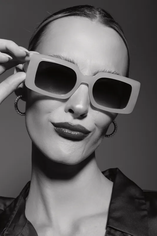 Confident white woman with pale skin adjusts large sunglasses in black and white studio portrait, fashion and style concept.