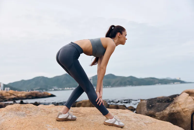 Young athletic woman stretching on rocks by the sea, wearing gray sportswear, showcasing determination and focus in a serene natural environment