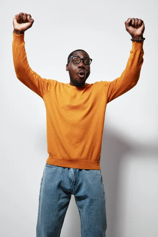 African American man with dark skin wearing orange sweater and glasses raising hands in excitement isolated on white background. Casual clothing and joyful expression.