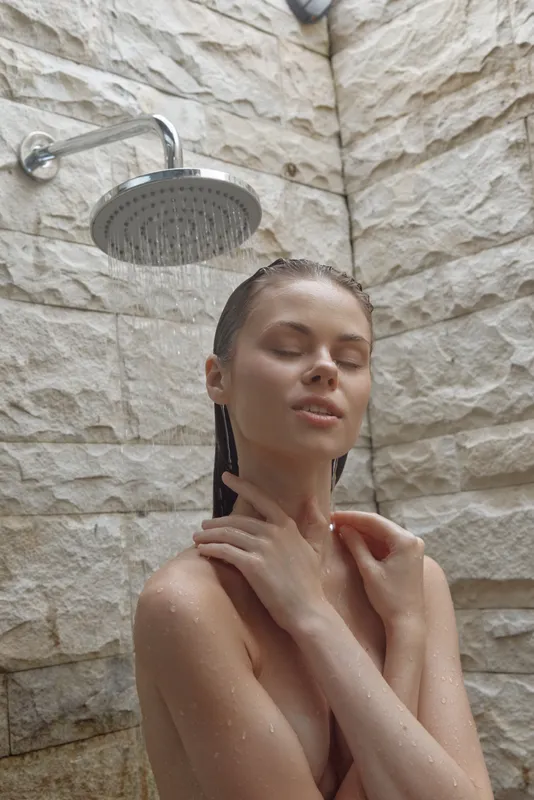 young woman enjoying outdoor shower with stone wall background, serene expression, natural skin, and soft water droplets creating a calming atmosphere