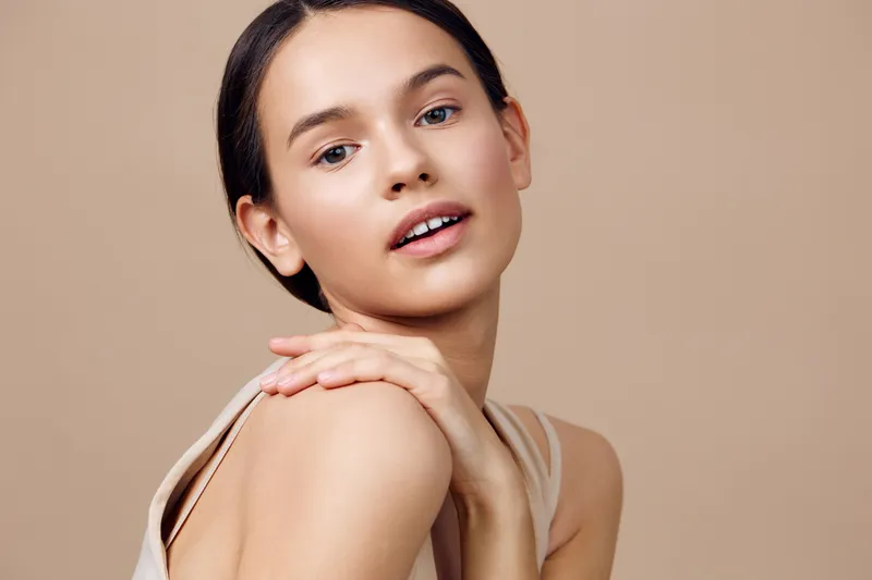 Young woman with a natural makeup look poses gracefully, showcasing radiant skin, soft expressions, and a minimalist style on a warm beige background