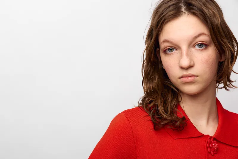 Young woman with wet brown hair and freckles wears red sweater with collar, looking serious. Studio portrait on white background with natural expression and soft lighting.