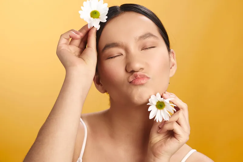 Asian woman with a flower near her head, eyes closed and lips puckered in a playful kiss, set against a warm orange background. The portrait conveys softness, charm, and calm energy.
