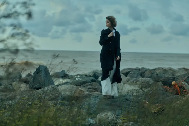 Outdoor fashion seascape on a windy rocky shore, coat and dress fluttering, capturing a moody windblown scene along the coast with dramatic silhouette, calm ocean and cloudy sky.