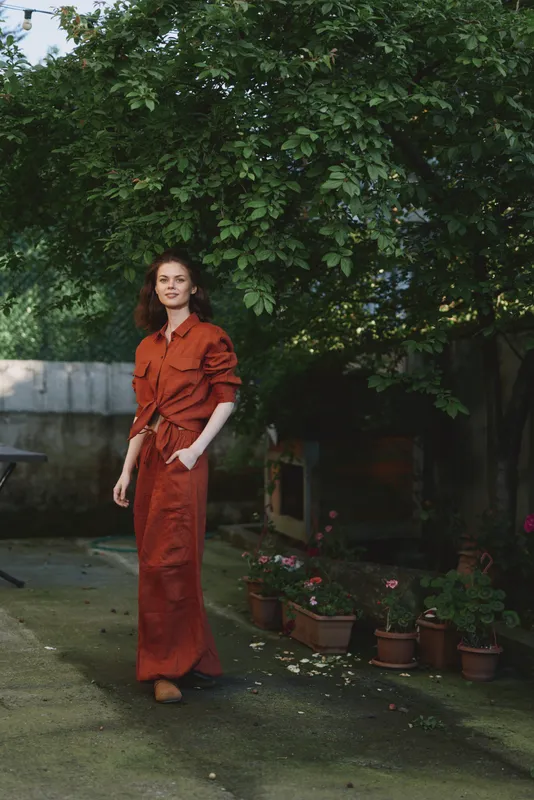 Woman in a rust dress standing outdoors in a lush garden, posing gracefully for a lifestyle portrait. Natural daylight creates a calm, casual, elegant mood