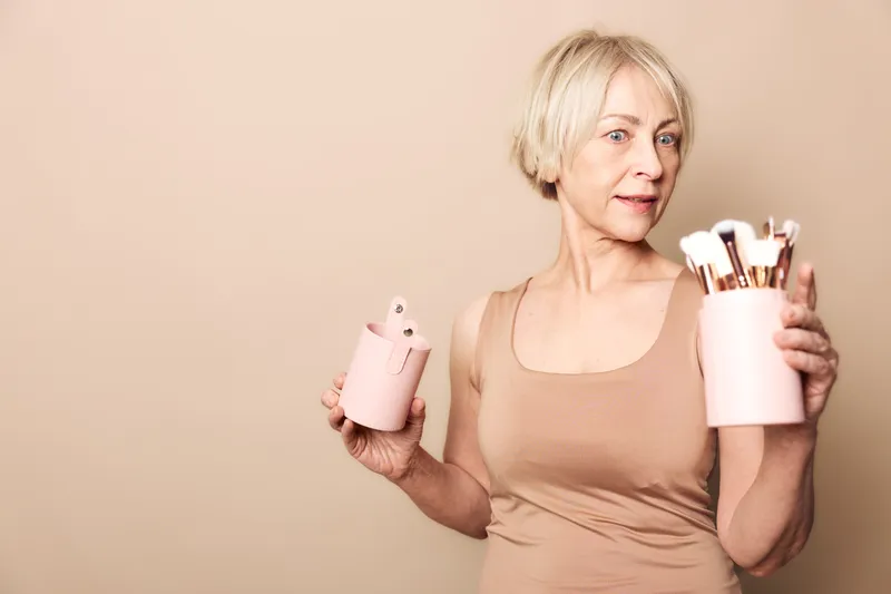 Mature woman with short blonde hair holds pink containers filled with makeup brushes, wearing a beige top against a soft neutral background. Concept of beauty routine and self-care.