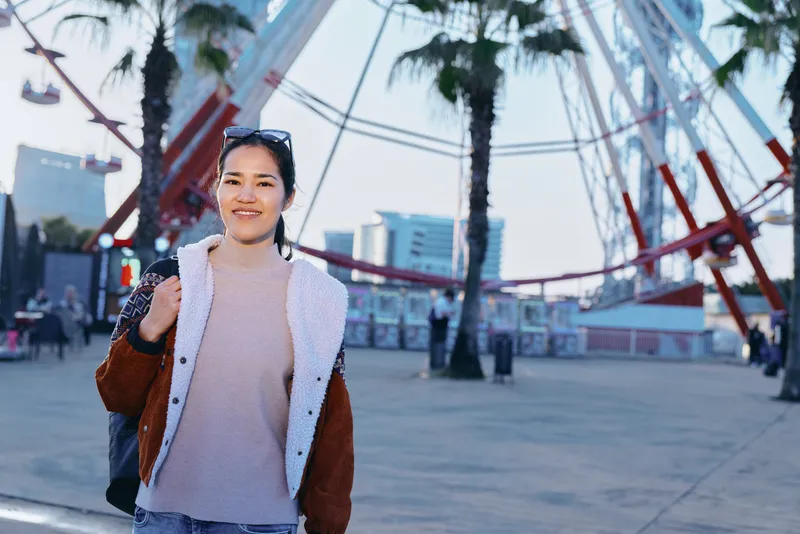 Woman in casual jacket and sweater smiles while strolling along a boardwalk with palm trees and a ferris wheel in the background, showcasing relaxed urban style and sunny travel mood