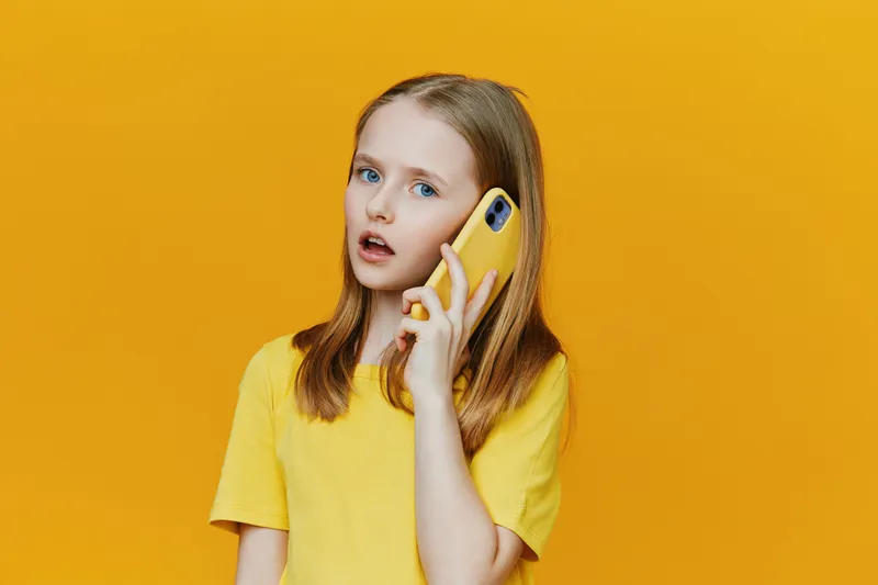 Young girl with blue eyes talking on a yellow phone, wearing a bright yellow t shirt against a vibrant yellow background, expressing curiosity and surprise