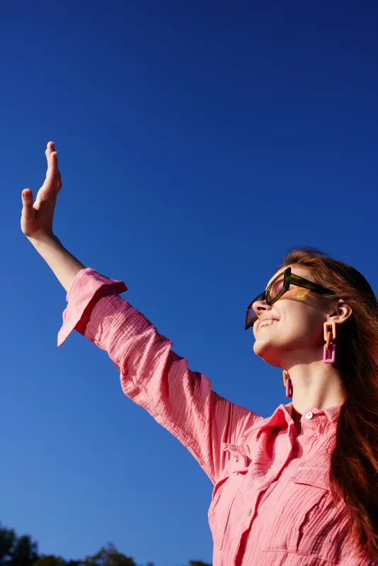 A joyful young woman with light brown hair, wearing a pink shirt and sunglasses, reaching out towards the clear blue sky exuding positivity and warmth