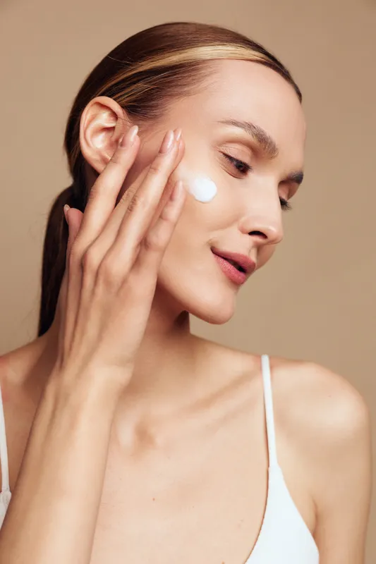 Young woman with clear skin applies moisturizer cream on her face with fingers, showing skincare routine and natural beauty, isolated on beige background with soft lighting.