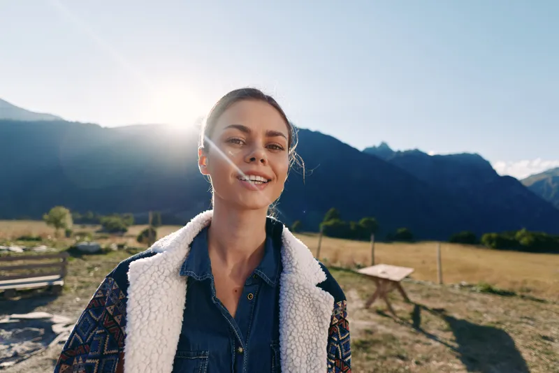 Woman portrait with smile against mountains outdoors at golden sunlight in countryside. Casual jacket and denim shirt, natural expression, travel lifestyle, scenic meadow background and leisure.
