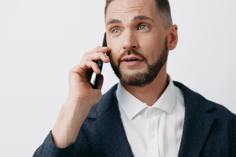 A professional man in business attire on a light blue solid background speaks on a mobile phone with focused emotion, showing calm confidence and strategic dialogue in a remote work moment.