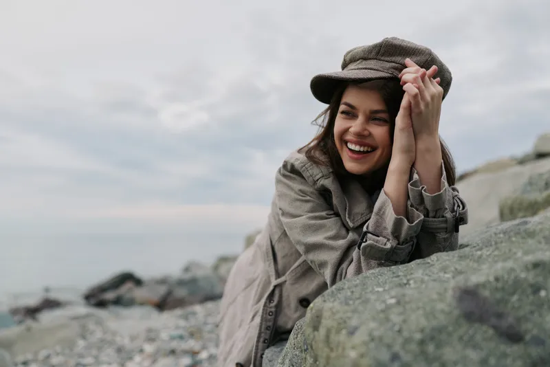 Smiling young woman in casual clothes and hat leans on rock near rocky beach under cloudy sky. Concept of happiness, nature, and relaxed outdoor lifestyle by the sea.