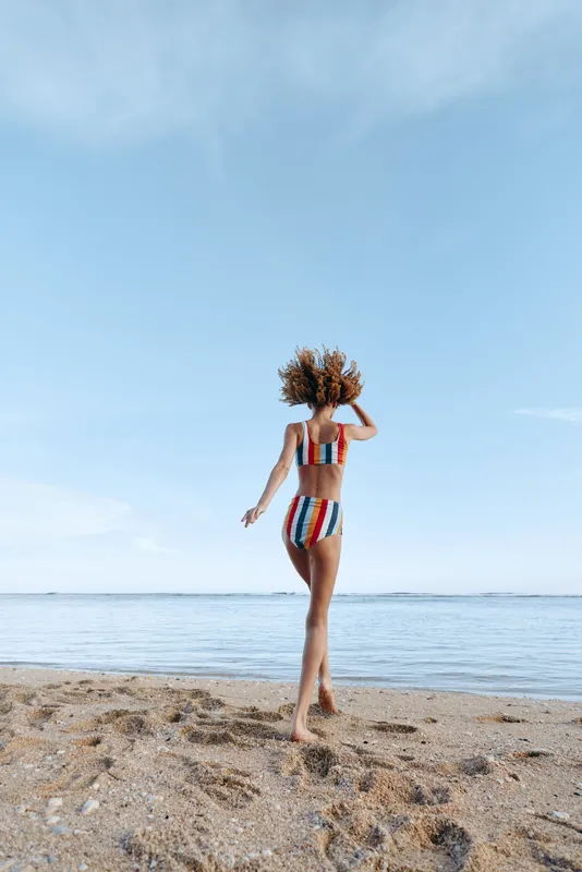 Woman walking on beach beside ocean under blue sky wearing striped swimsuit, summer travel mood, back view, natural sunlight, calm waves in distance, lifestyle vacation, calm horizon