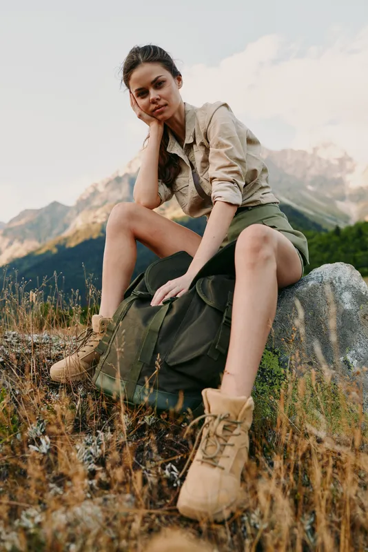 Young woman traveler sits on rock in mountain field wears khaki shorts and jacket with boots looks thoughtfully enjoying outdoor lifestyle adventure in nature.