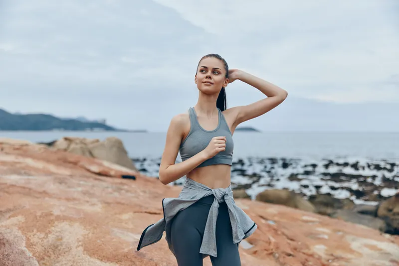 Fit young woman jogging on rocky beach, wearing gray sportswear, with a confident smile, overcast sky, promoting healthy lifestyle and fitness