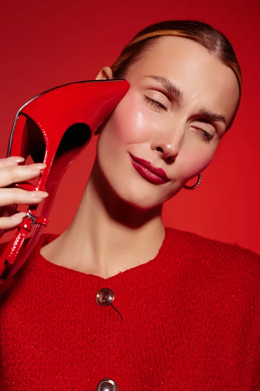 Close-up of a smiling woman in red sweater holding a red high heel shoe near her face on red background. Concept of fashion, style, beauty, and confident expression.