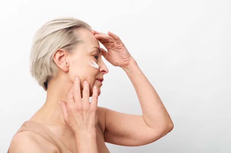 Mature woman with short blonde hair applies white face cream on cheek and forehead. Skincare routine concept for aging skin, beauty care, and self-care with natural light on white background.