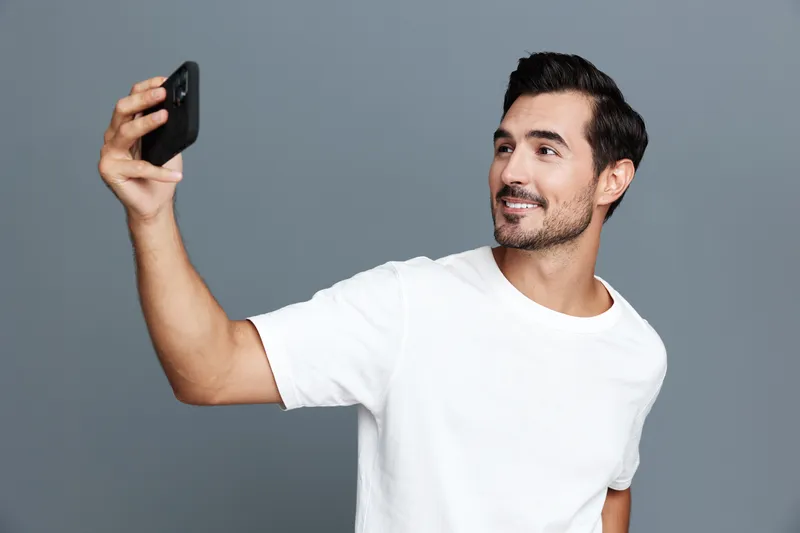 Smiling man taking selfie with smartphone, casual white t-shirt, standing against gray background, showcasing happiness and modern lifestyle in studio setting.