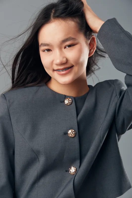 Young Asian woman smiling with braces wearing grey jacket and touching hair in studio. Portrait of happy female model with natural expression and loose long black hair on neutral background.