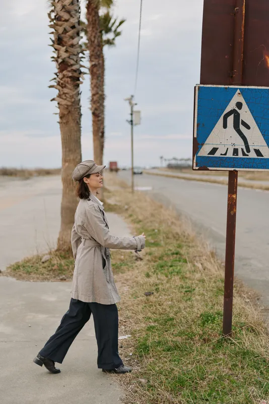 Young woman wearing coat and cap walking near pedestrian crossing sign on roadside with palm trees and cloudy sky in background. Concept for urban lifestyle and street crossing safety.