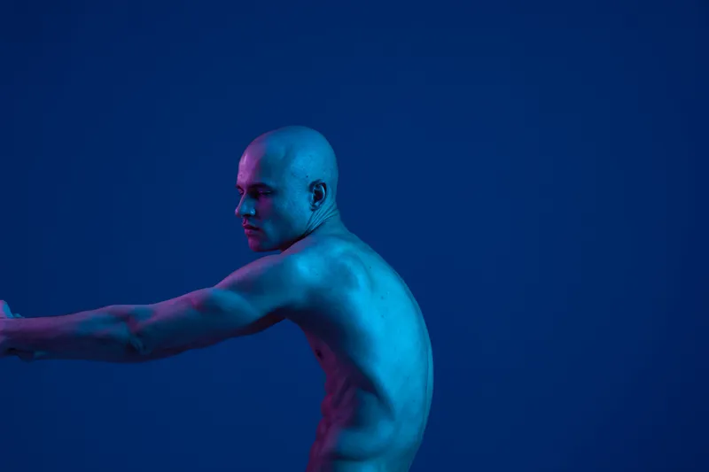 Bald man posing gracefully with outstretched arms, showcasing artistic expression in a captivating blue light. This image highlights the beauty of human form and emotion. ###