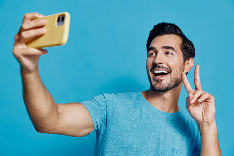 Smiling man taking selfie with mobile phone on vibrant blue background, showing peace sign with hand, enjoying moment of happiness and connection.
