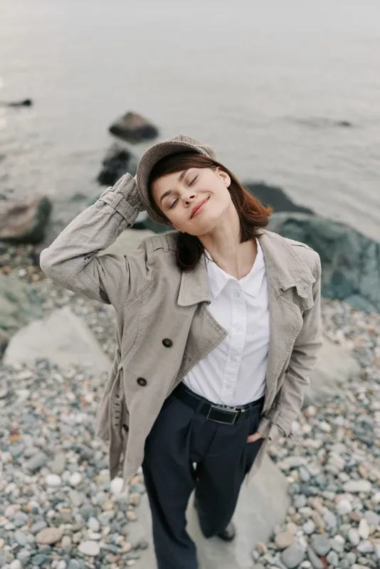 Young woman in casual jacket and cap stands on rocky beach with eyes closed, smiling gently. Relaxed mood and connection with nature by the water shore on overcast day.