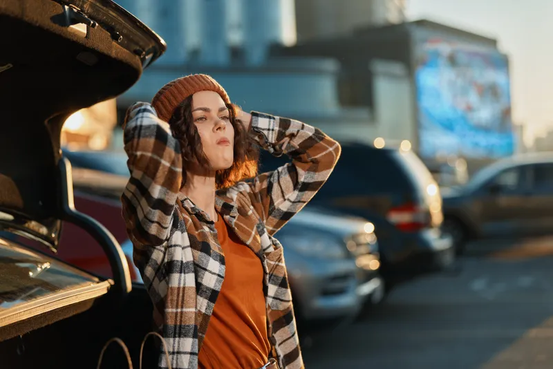 Woman leans against car trunk in parking lot at sunset, wearing beanie and plaid shirt, relaxed pose conveying authenticity, candid lifestyle and golden hour glow with mindful living mood.