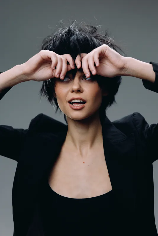 Smiling woman with short black hair wearing a black jacket on gray background expressing playful emotions