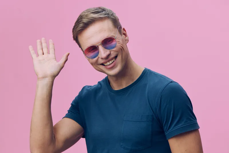 Smiling man wearing stylish sunglasses and a blue t-shirt, waving hand and looking at the camera against a pink background. Studio portrait of a cheerful young male with casual style, upbeat mood, and