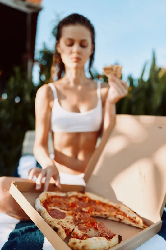 A relaxed woman in a white top sits outdoors with an open pizza box, savoring a slice under bright sun, capturing casual outdoor dining and a simple lifestyle moment.