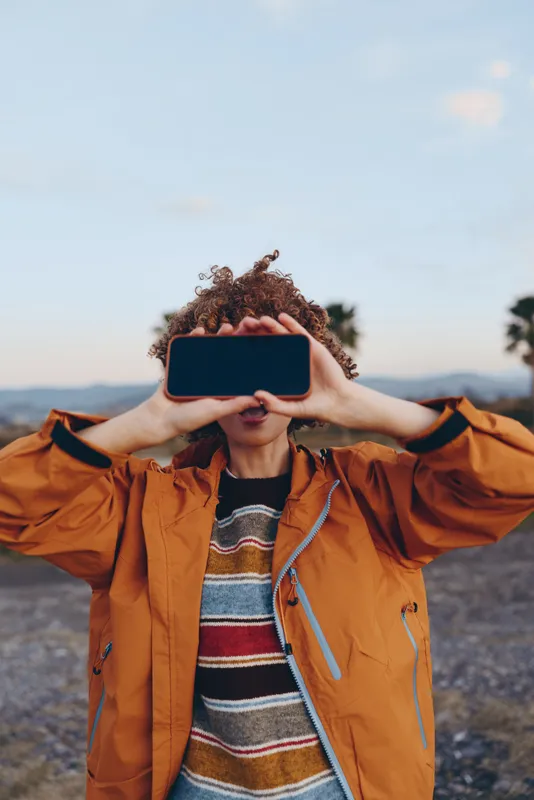 Woman lifestyle outdoors, curly hair, orange jacket, holding smartphone up to cover face, looking toward camera, scenic mountain background, copy space, casual travel moment, modern digital