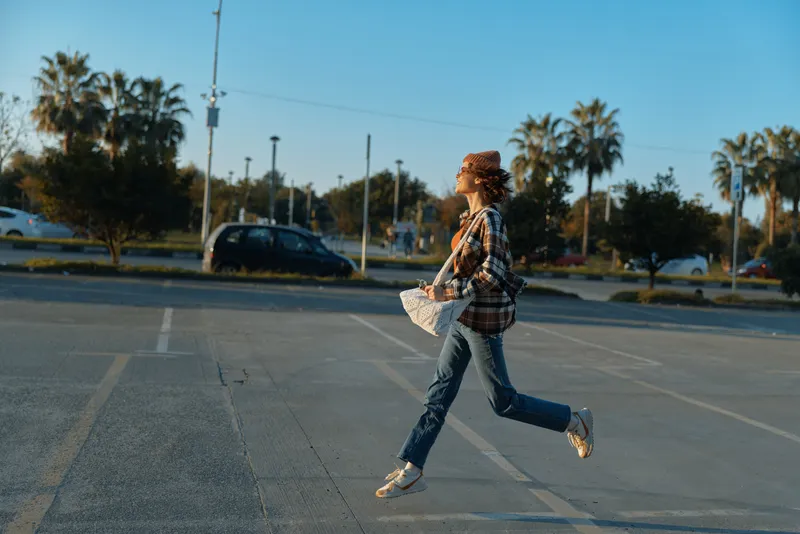 Woman running across empty parking lot at golden hour, joyful motion and casual style with tote bag, urban candid lifestyle portrait emphasizing authenticity and golden hour glow.