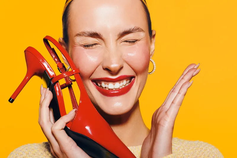 Happy woman with red lipstick holds red high heel shoe near face, eyes closed and smiling. Bright yellow background creates cheerful mood. Concept of fashion, beauty, and joy.