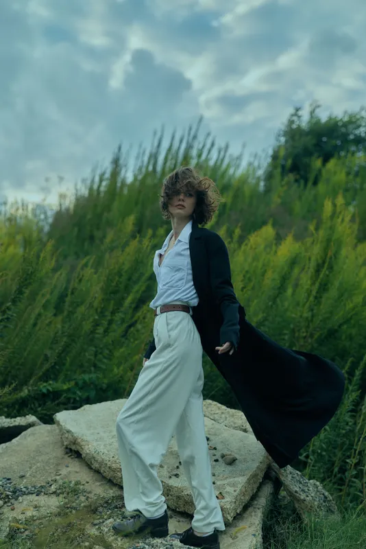 Fashion model outdoors wearing white suit with flowing black coat, poised on rocks among green plants, wind swept movement creating dramatic elegance and energy.