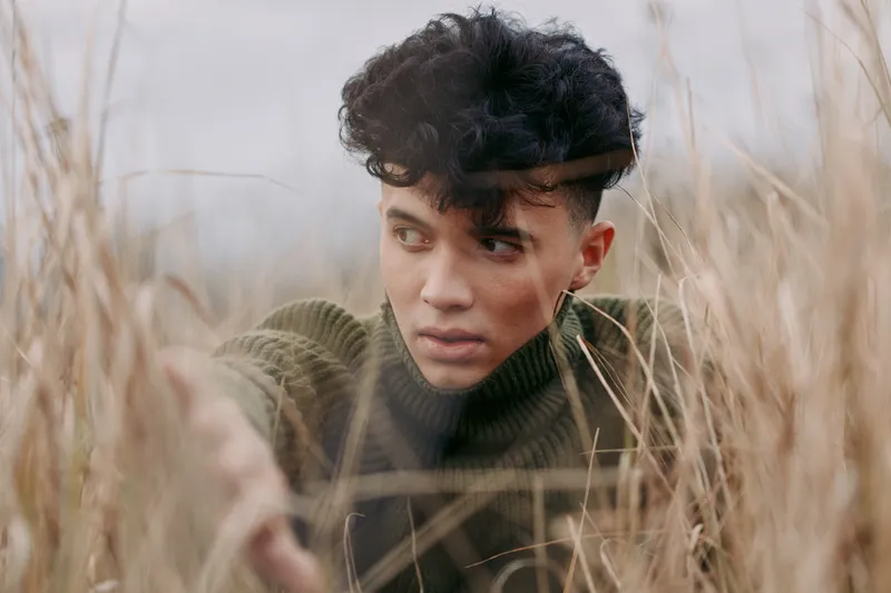 A candid portrait of a young man peering through tall dry grass, captured with natural light and subtle tones to convey authenticity and quiet strength in a real moment.
