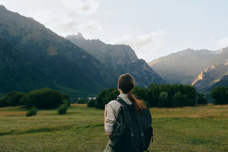Woman traveler with backpack stands in green valley looking at mountain range under cloudy sky, enjoying nature and outdoor lifestyle during daytime.