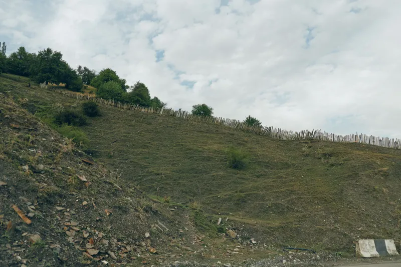 Hillside with rocky slope and sparse vegetation under a partly cloudy sky, showcasing rugged terrain and vast natural landscape with earthy textures