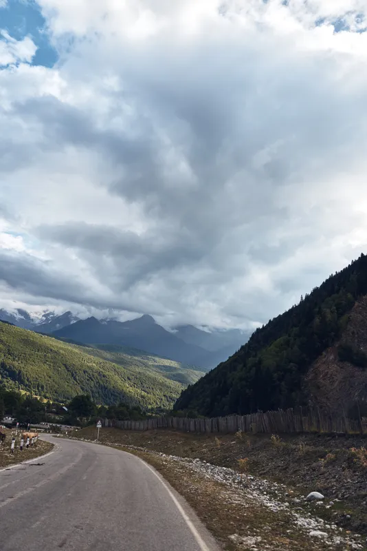 A winding road cuts through a rugged mountain valley under a dramatic cloudy sky, offering a scenic landscape that invites exploration and peaceful travel
