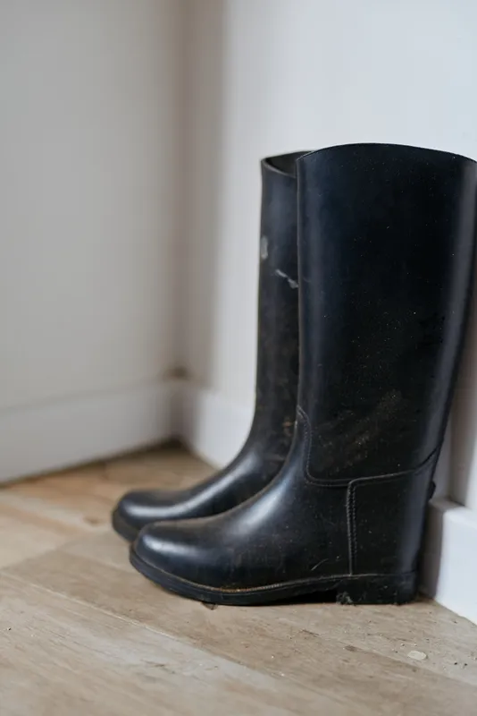 A pair of black rubber boots rests beside a white wall on a wooden floor, suggesting ready outdoor wear in a simple home scene with quiet, everyday routine and anticipation
