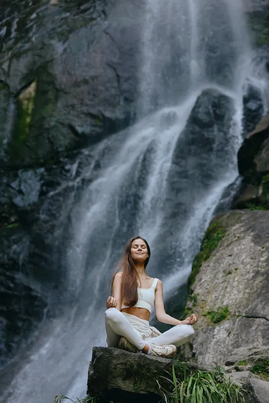 Calm woman meditating outdoors near a waterfall, wearing white clothes, serene expression, natural rocks and green plants surrounding, peaceful nature setting.