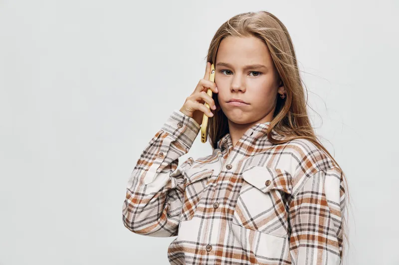 Girl with long hair wearing plaid shirt talking on smartphone with serious expression isolated on white background. Child communication, technology and portrait concept.