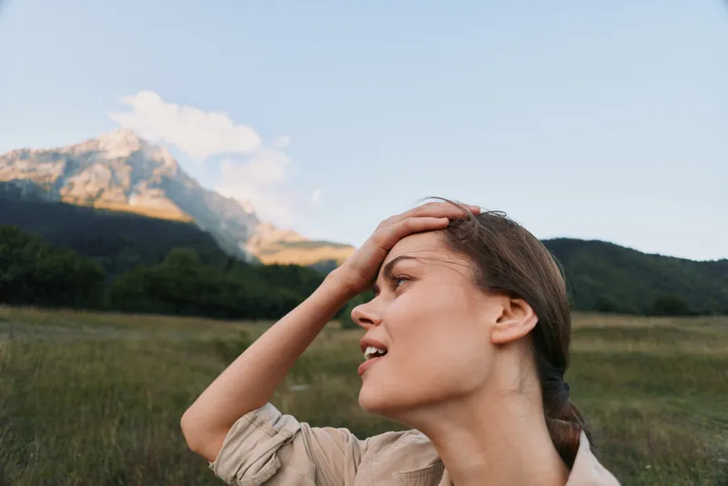 Young woman with brown hair enjoys a sunny day outdoors with mountains in the background while touching her forehead and smiling softly.