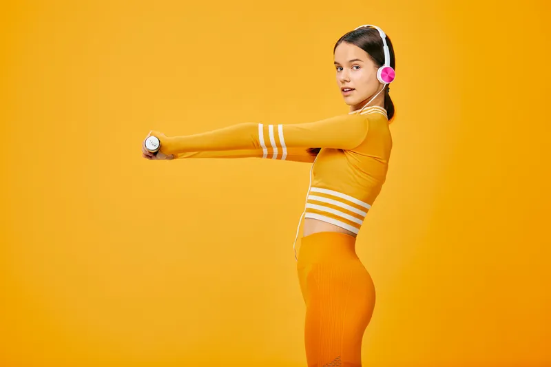Fitness woman in sporty orange outfit performing a stretch with outstretched arms against a vibrant orange backdrop, wearing headphones, conveying energy, health, and an active lifestyle in a studio
