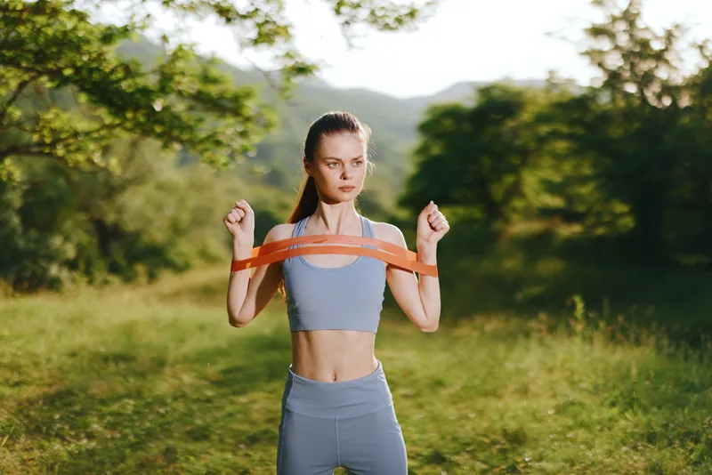 Young athletic woman exercising outdoors with resistance bands in a natural green setting, embodying a healthy lifestyle and fitness motivation
