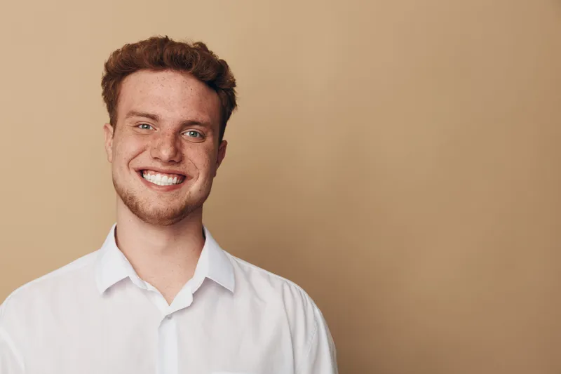 Smiling young man with curly hair wearing white shirt stands against beige background. Portrait of happy confident male model with natural expression and casual style in studio lighting.